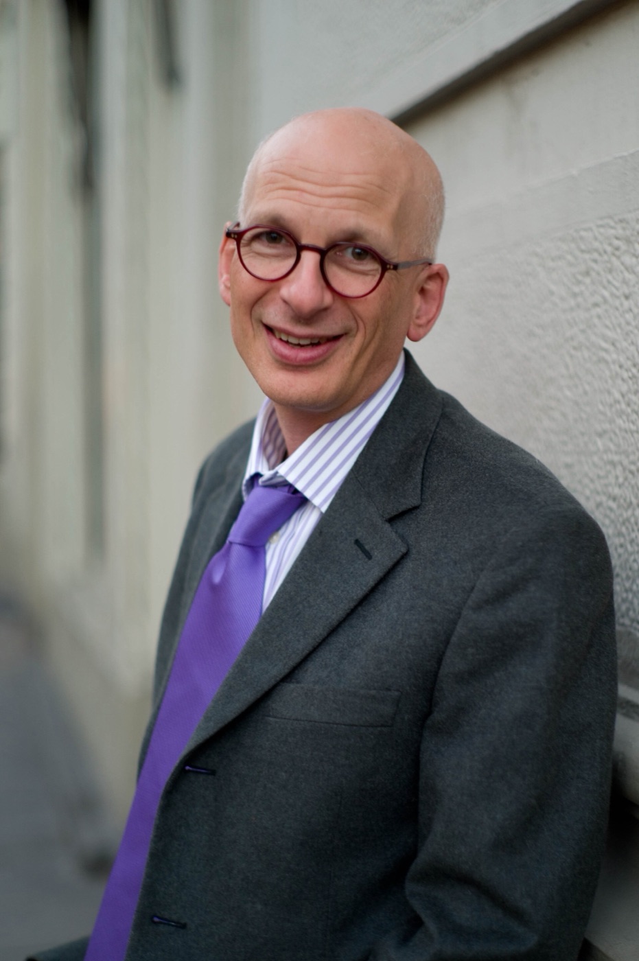 Seth Godin, de óculos redondos e gravata roxa, sorrindo encostado numa parede clara
